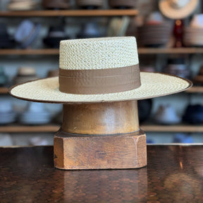 Canero Bolero straw hat with wide brim and brown band displayed on a wooden hat stand, set against a blurred background of various hats and accessories.