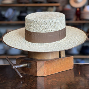 Canero Bolero straw hat with a wide brim and brown fabric band, displayed on a wooden hat block in a shop setting with other hats in the background.
