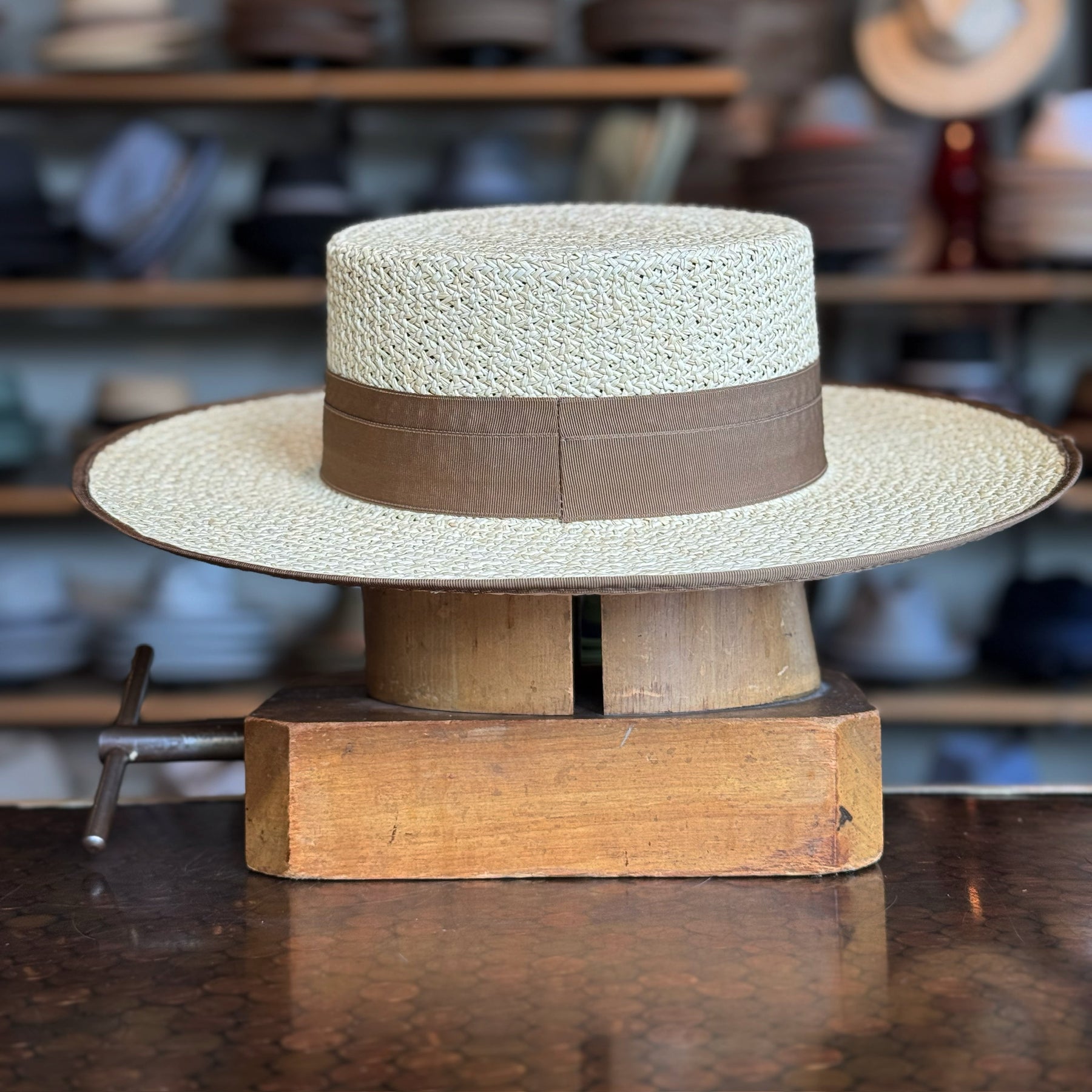 Canero Bolero straw hat with a wide flat brim and a brown fabric band displayed on a wooden hat block in a shop setting.