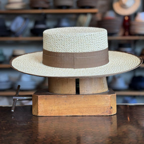 Canero Bolero straw hat with a wide flat brim and a brown fabric band displayed on a wooden hat block in a shop setting.