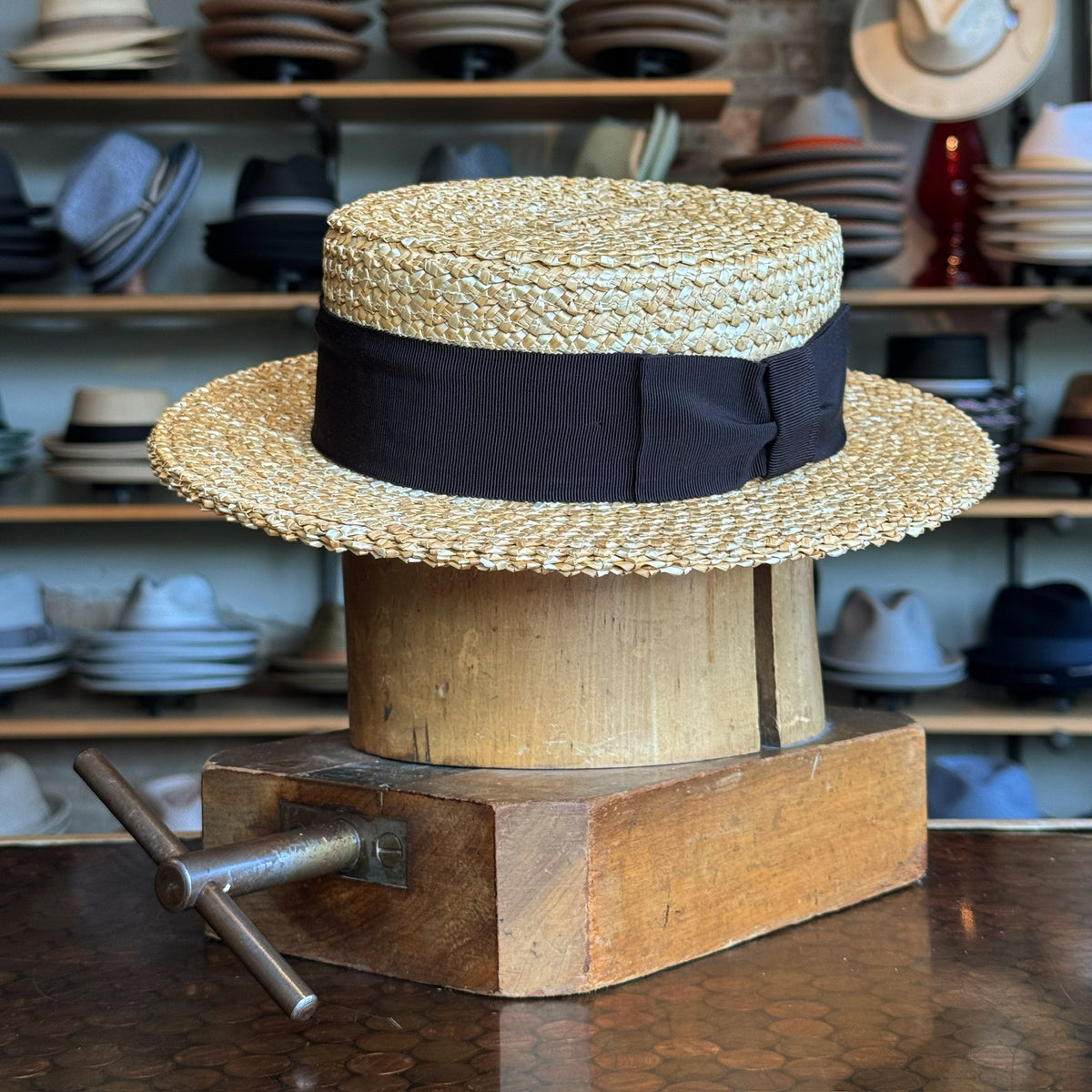 Canoutier Boater straw hat with a wide black ribbon band, displayed on a wooden hat block in a shop filled with various hats in the background.