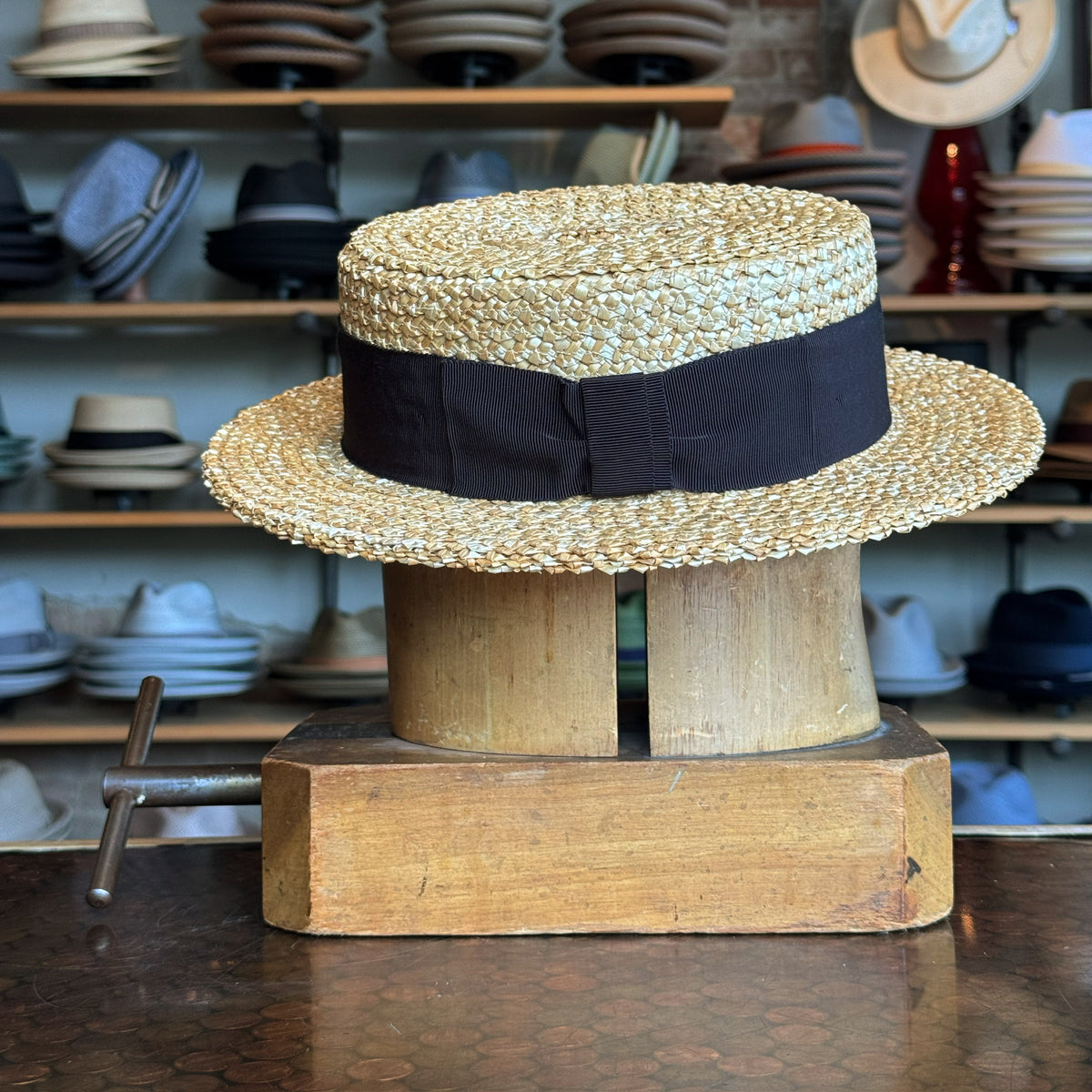 Canoutier Boater straw hat with a wide black ribbon band displayed on a wooden hat block in a shop filled with various hats in the background
