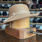 Caramel Mandy Wide Brim Cloche Hat displayed on a wooden hat block, featuring a textured caramel color and a decorative stitched band, surrounded by various hats in the background.