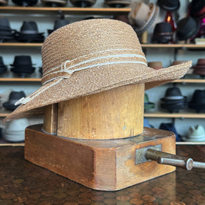 Caramel Mandy Wide Brim Cloche Hat displayed on a wooden hat block, featuring a textured caramel color and a decorative stitched band, surrounded by various hats in the background.