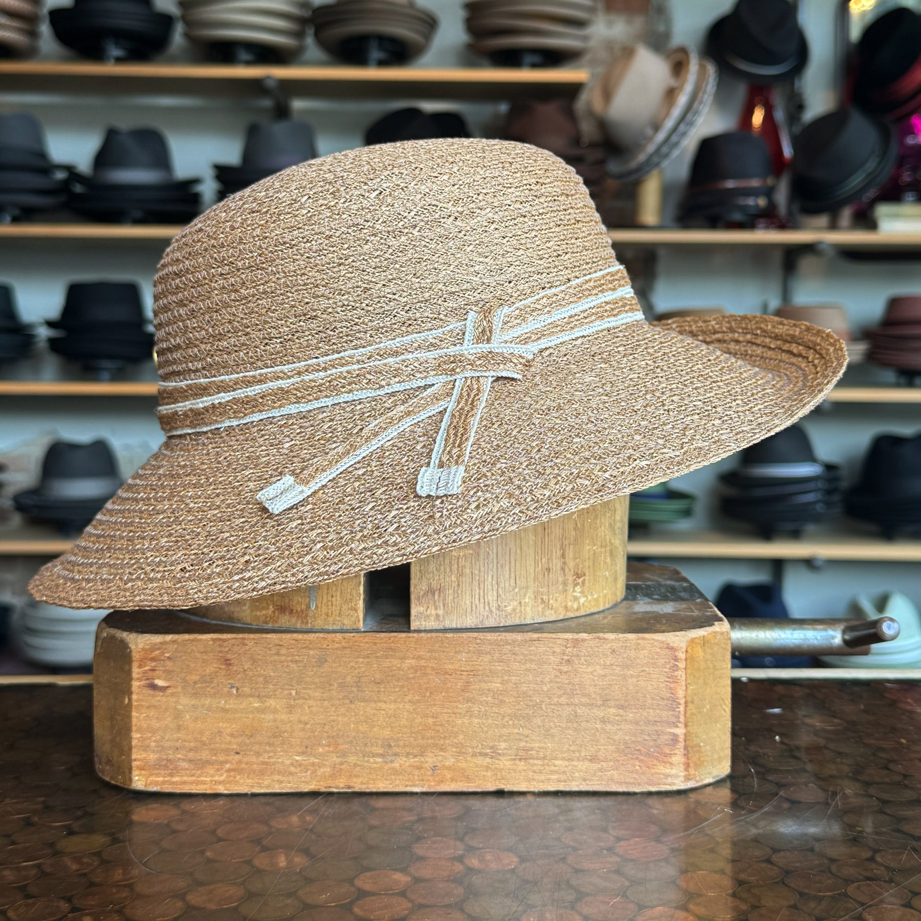 Caramel Mandy Wide Brim Cloche Hat in a woven straw design with a decorative band detail, displayed against a backdrop of various hats on shelves.