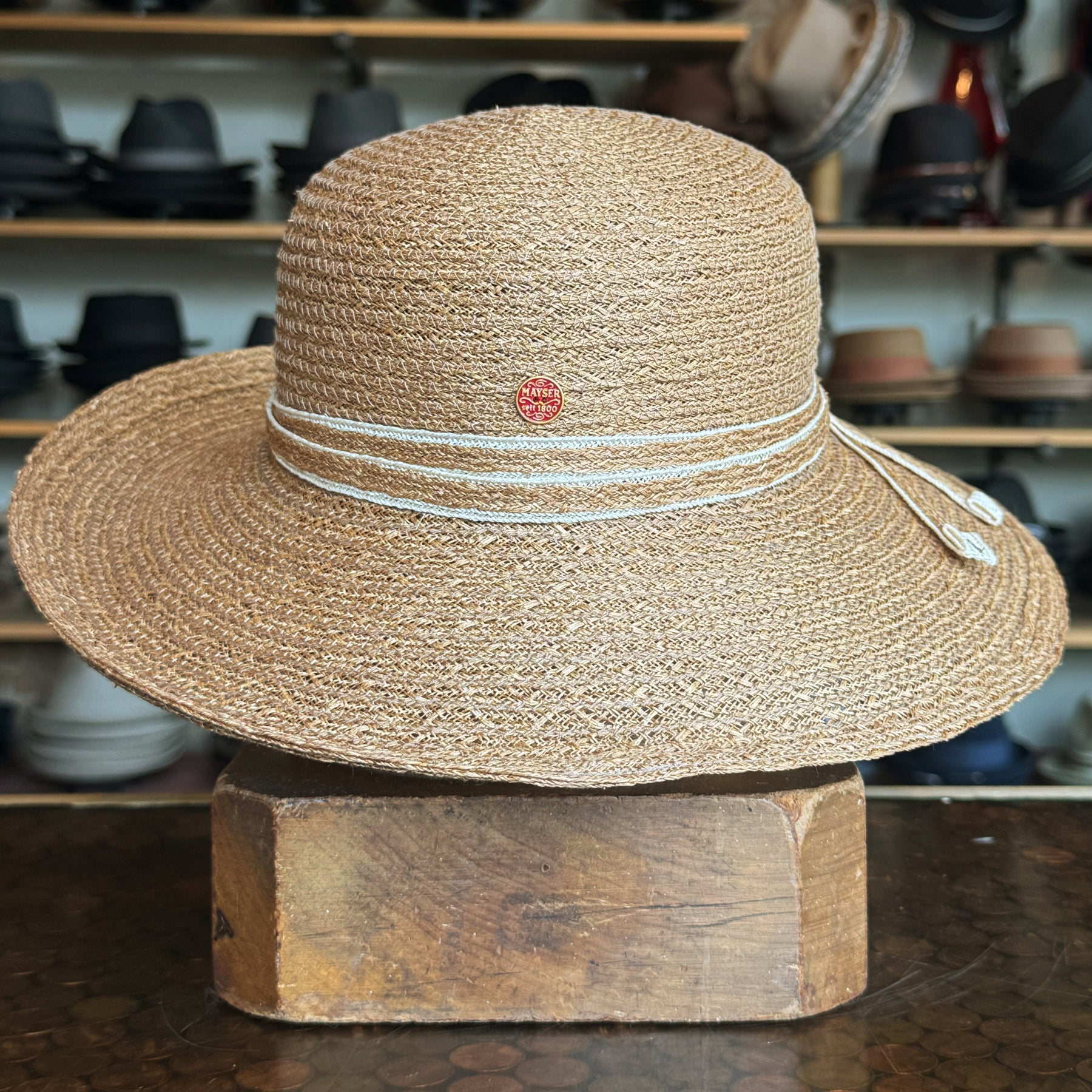Caramel Mandy Wide Brim Cloche Hat made of woven material with decorative white cord detail and small round emblem, displayed on wooden block with hats in background.