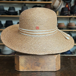 Caramel Mandy Wide Brim Cloche Hat made of woven material with decorative white cord detail and small round emblem, displayed on wooden block with hats in background.