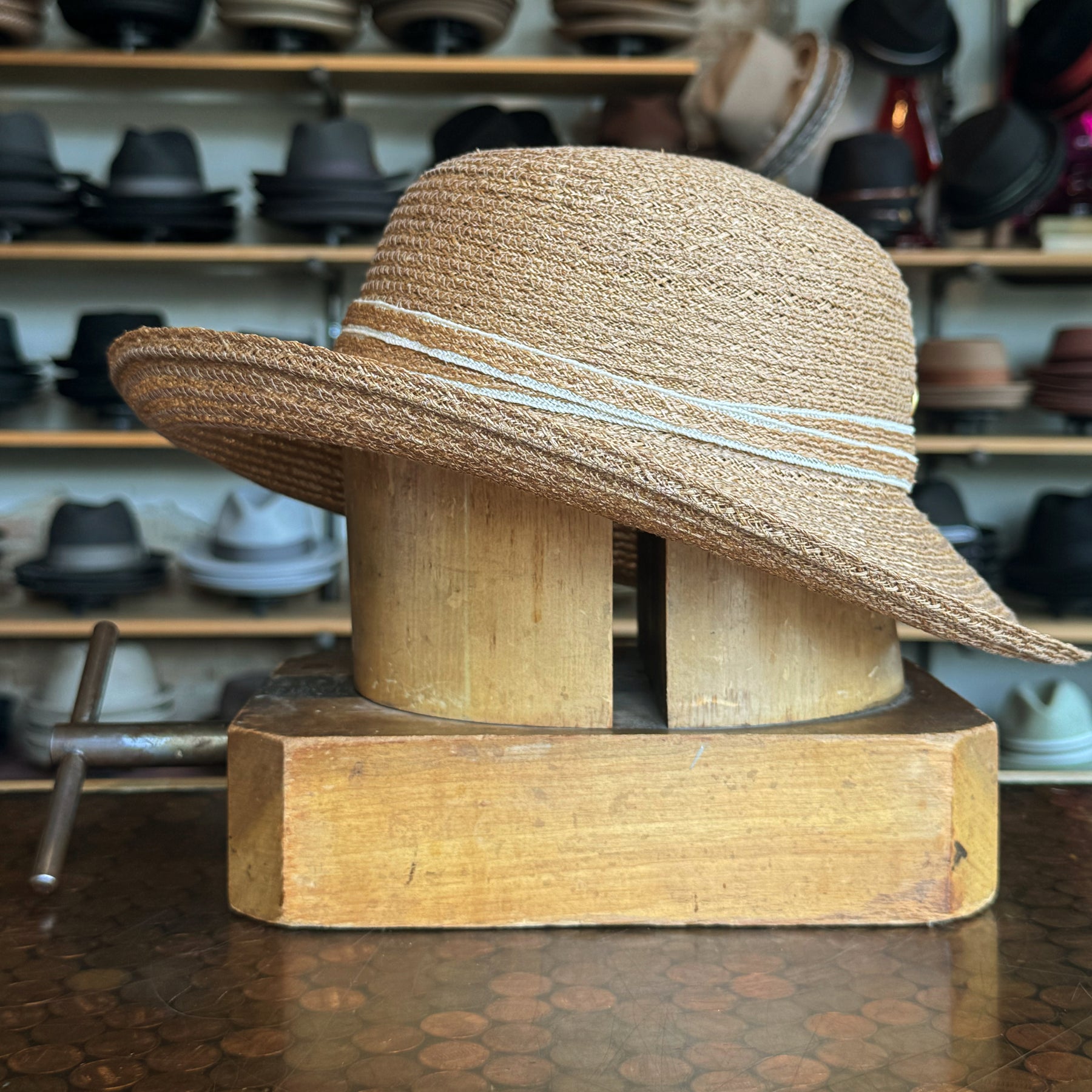 Caramel Mandy Wide Brim Cloche Hat displayed on a wooden hat block with a textured woven design and subtle decorative stripes, surrounded by various hats in the background.