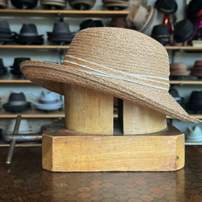 Caramel Mandy Wide Brim Cloche Hat displayed on a wooden hat block with a textured woven design and subtle decorative stripes, surrounded by various hats in the background.