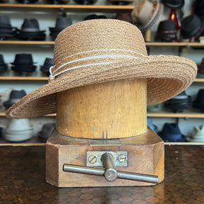 Caramel Mandy Wide Brim Cloche Hat displayed on a wooden hat block, showcasing its woven texture and light brown caramel color in a hat shop setting.