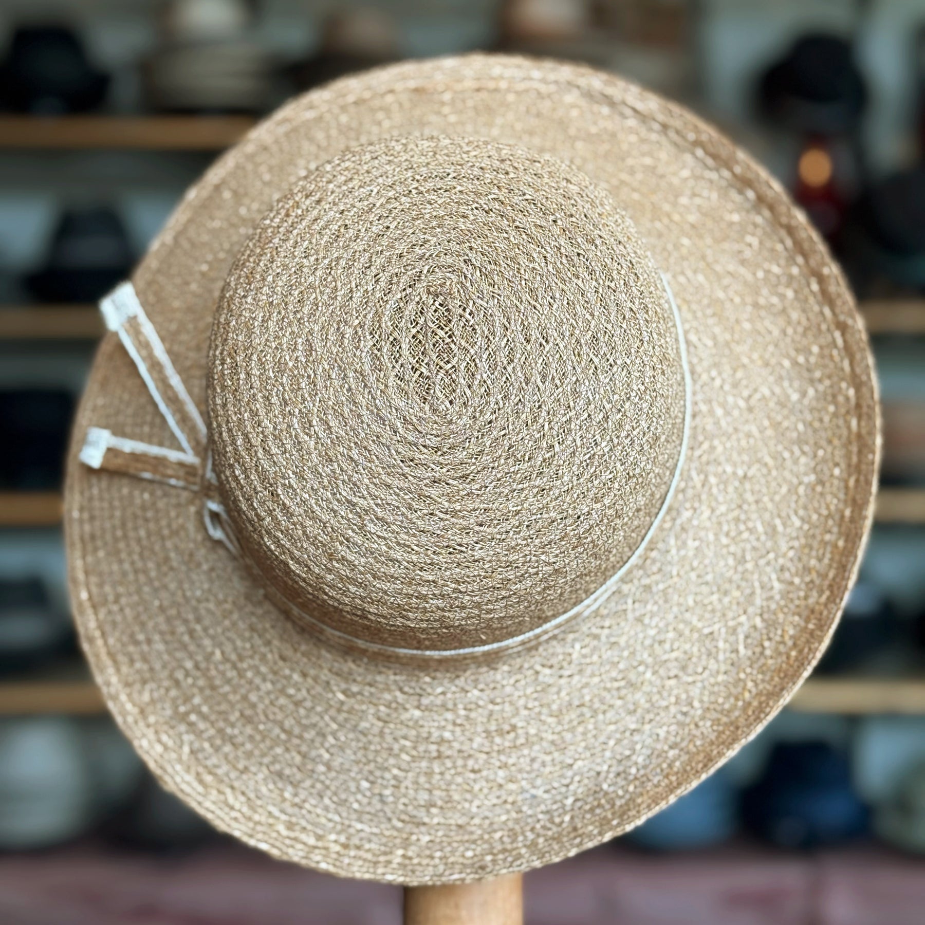 Caramel Mandy Wide Brim Cloche Hat made of woven natural fibers with a wide brim and decorative ribbon detail, displayed on a wooden stand in a hat shop.