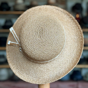 Caramel Mandy Wide Brim Cloche Hat made of woven natural fibers with a wide brim and decorative ribbon detail, displayed on a wooden stand in a hat shop.