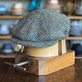 Classic Herringbone Newsboy cap in a textured herringbone pattern with subtle flecks of color, displayed on a wooden hat block in a hat shop setting.