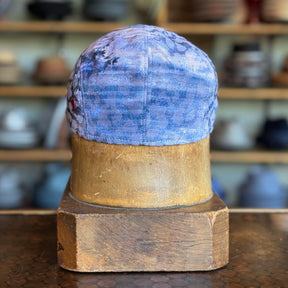 Back view of the Cloud Leopard 2-Panel Ivy Cap in a muted blue fabric with subtle leopard print, displayed on a wooden hat block in a hat shop background.