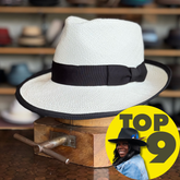 Clover Club white textured fedora hat with a black band and trim, displayed on a wooden stand in a store setting with various hats blurred in the background.