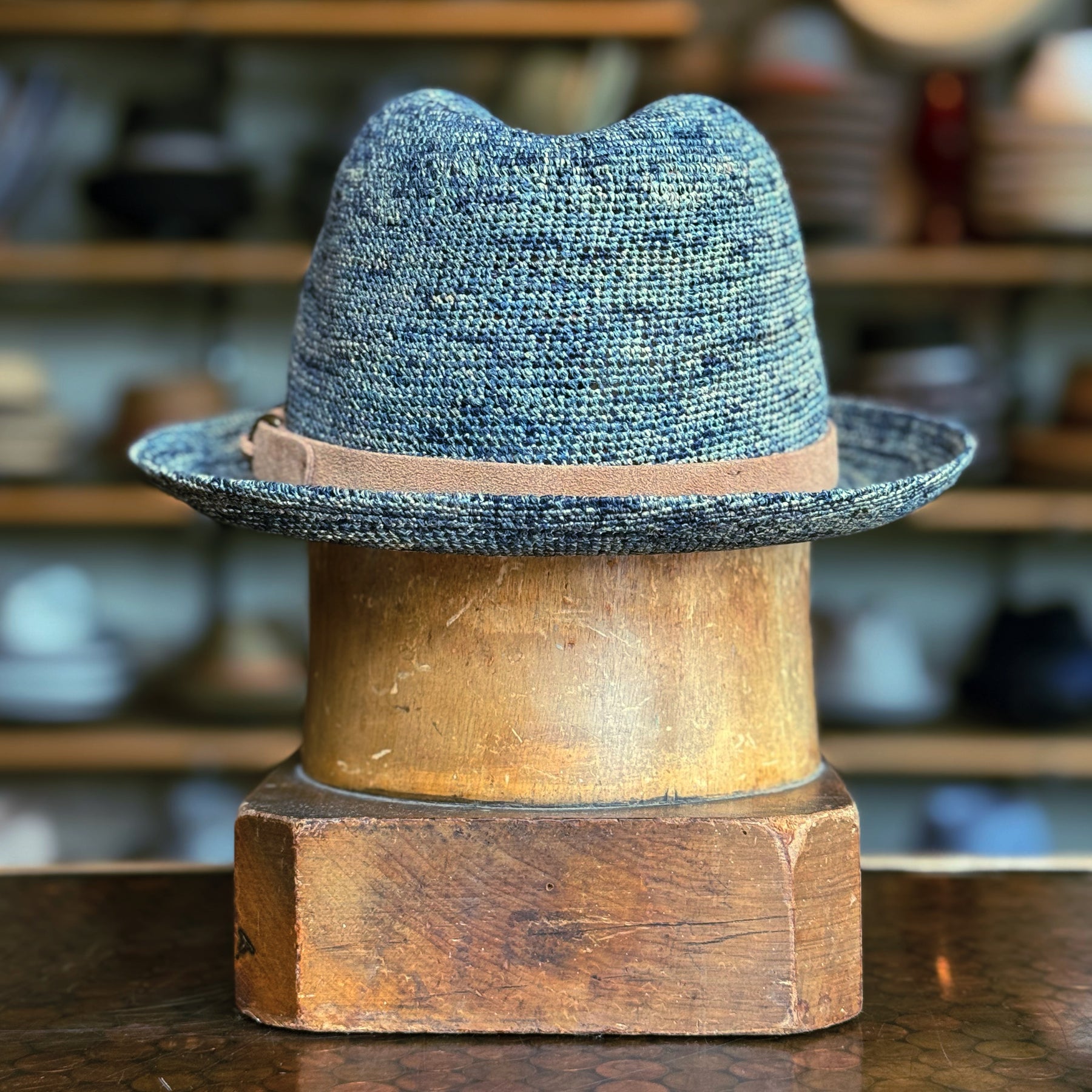 Back of Crochet Tino Fedora in a textured blue braided material with a light brown leather band, displayed on a wooden hat stand in a rustic setting.