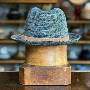 Back of Crochet Tino Fedora in a textured blue braided material with a light brown leather band, displayed on a wooden hat stand in a rustic setting.
