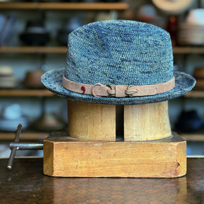 Side of Crochet Tino Fedora in a textured blue crochet material with a brown leather  band and buckle detail, displayed on a wooden hat block in a cozy workshop setting.