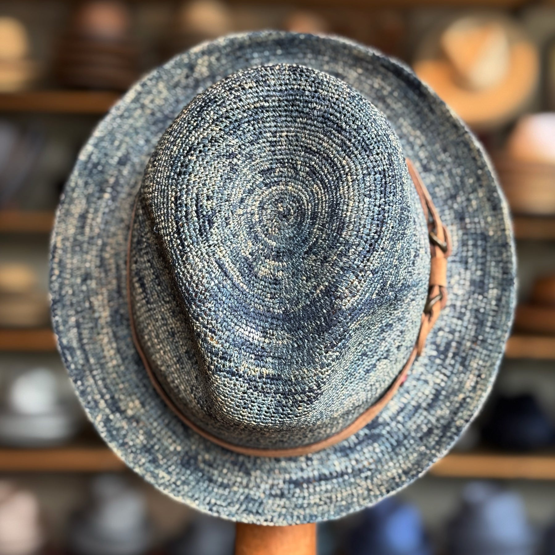 Top of Crochet Tino Fedora in a blue crochet design with a brown leather band, displayed on a wooden stand with blurred shelves of hats in the background.