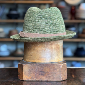 Back of Crochet Tino Fedora in green textured fabric with a brown leather  band, displayed on a wooden hat stand, showcasing a classic fedora shape and brim style.
