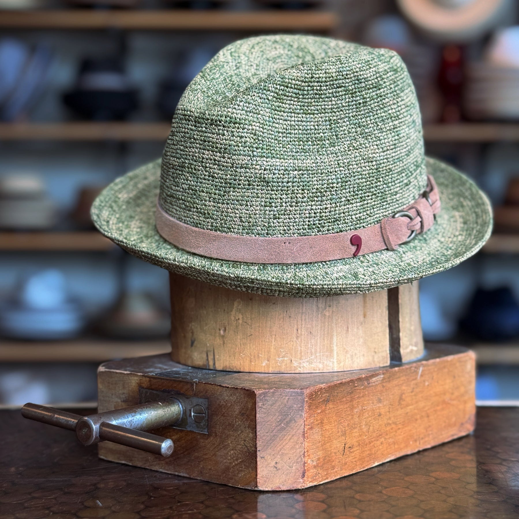 Crochet Tino Fedora in green with a brown leather  band and buckle detail, displayed on a wooden hat block in a shop setting with blurred shelves in the background.