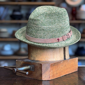 Crochet Tino Fedora in green with a brown leather  band and buckle detail, displayed on a wooden hat block in a shop setting with blurred shelves in the background.