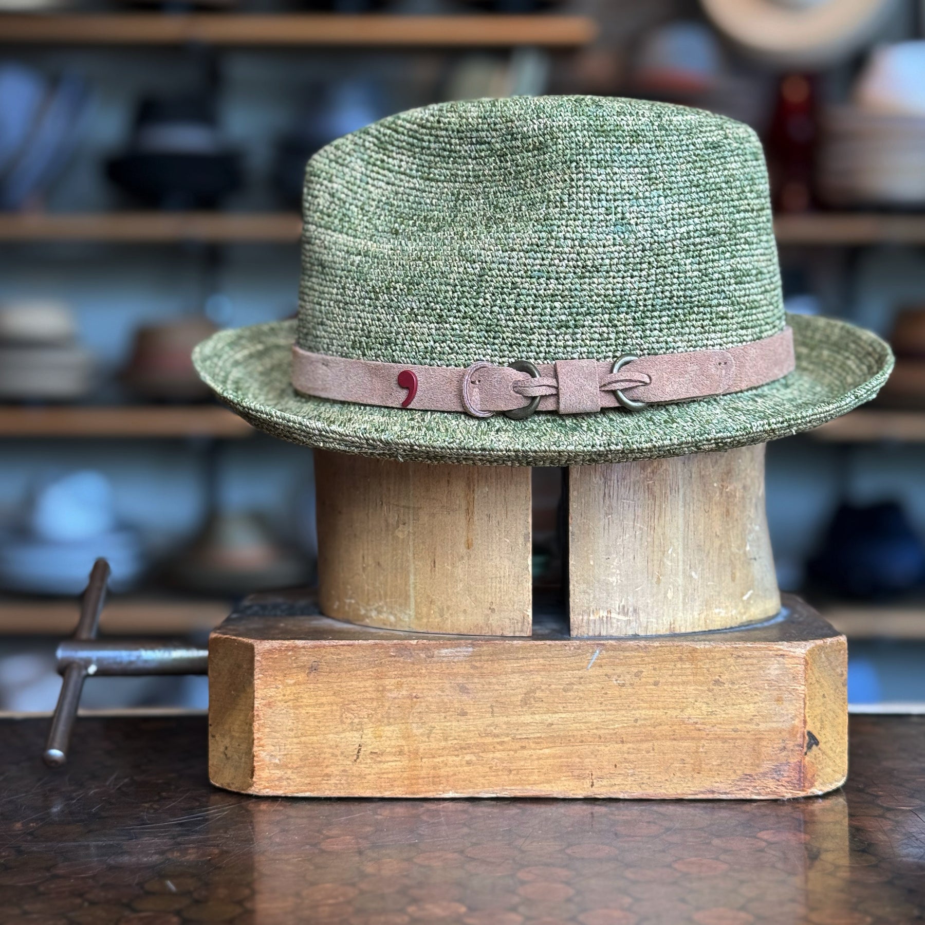 Side of Crochet Tino Fedora in a muted green shade with a brown leather  band, displayed on a wooden hat block in a cozy shop setting with shelves of accessories blurred in the background.