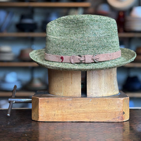 Side of Crochet Tino Fedora in a muted green shade with a brown leather  band, displayed on a wooden hat block in a cozy shop setting with shelves of accessories blurred in the background.