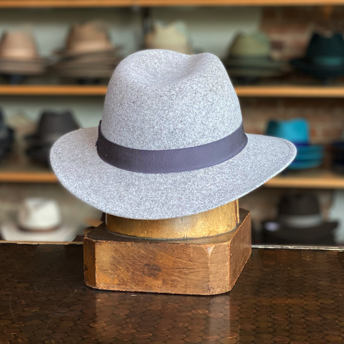Curtis flat cap in speckled light gray fabric with a dark band, displayed on a vintage wooden stand inside a hat shop with various hats on shelves in the background.