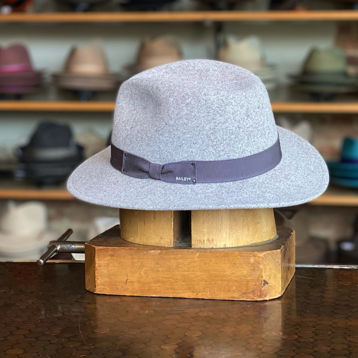 Curtis gray felt fedora hat with a black ribbon band displayed on a wooden hat block in a shop setting with blurred hats in the background.