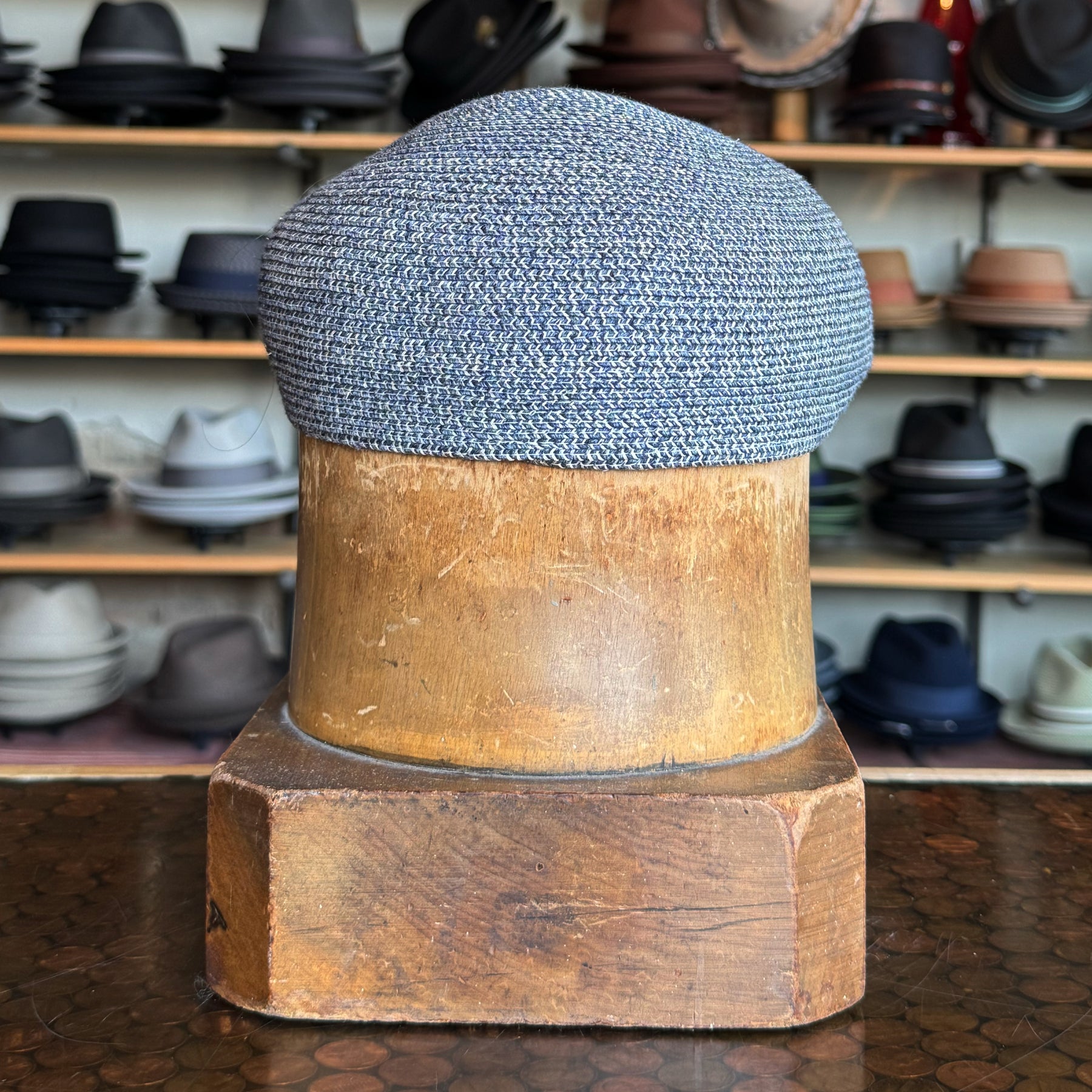 Dan Ascot Cap in textured blue fabric displayed on a wooden hat block with various hats on shelves in the background.