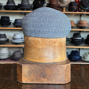 Dan Ascot Cap in textured blue fabric displayed on a wooden hat block with various hats on shelves in the background.