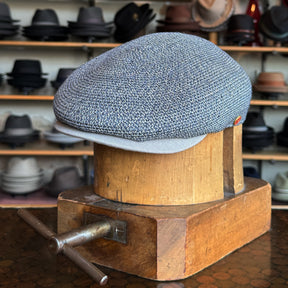 Dan Ascot Cap in textured denim fabric displayed on a wooden hat block with various hats blurred in the background, showcasing a stylish flat cap accessory.