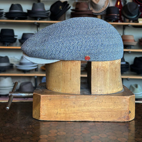 Dan Ascot Cap in a textured blue fabric displayed on a wooden hat block with additional hats visible in the background, showcasing a classic newsboy style accessory.
