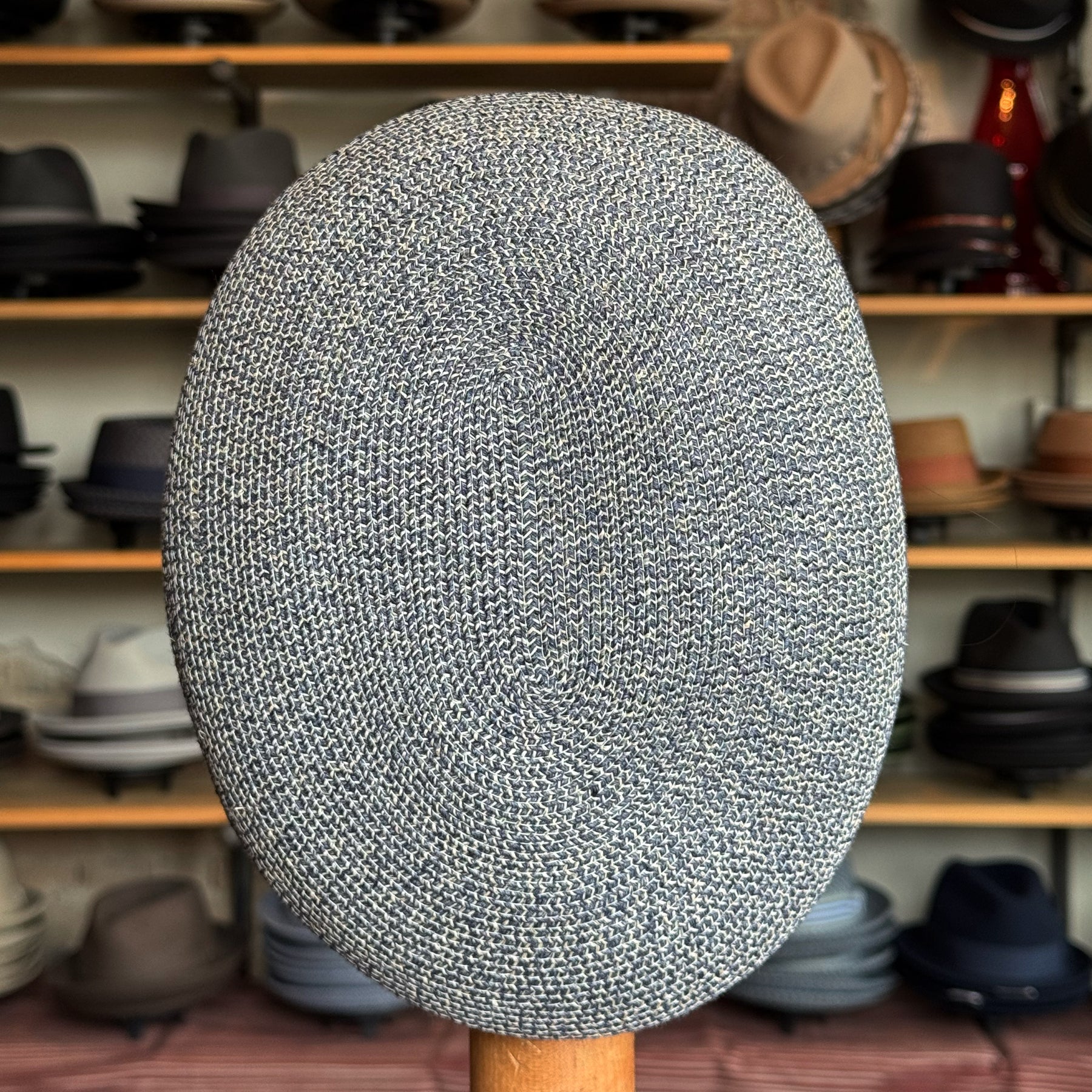 Dan Ascot Cap in a textured blue and white woven fabric, displayed front-facing on a wooden stand with a variety of hats blurred in the background.