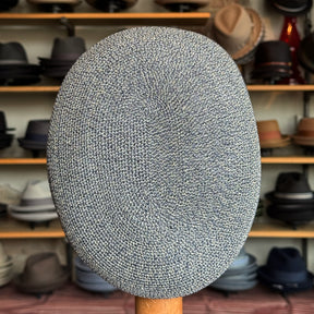 Dan Ascot Cap in a textured blue and white woven fabric, displayed front-facing on a wooden stand with a variety of hats blurred in the background.