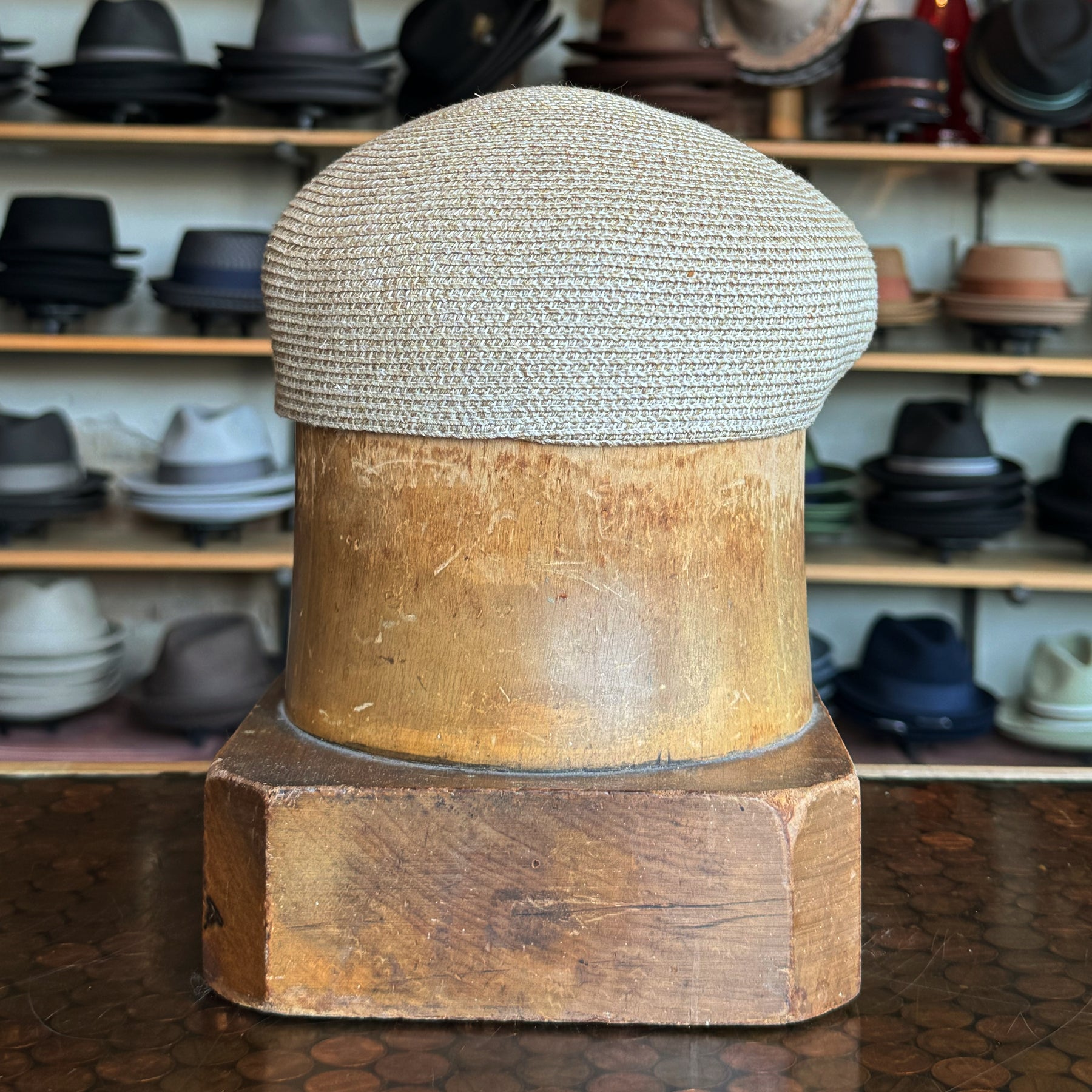 Natural beige woven Dan Ascot Cap displayed on a wooden hat block with shelves of various hats in the background, showcasing its classic newsboy style.