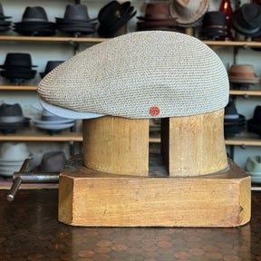 Side view of a natural-colored woven Dan Ascot Cap displayed on a wooden hat block with various hats visible on shelves in the background.