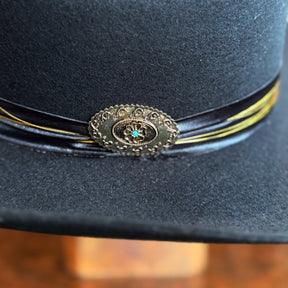 Close-up of a dark felt brim hat with a decorative vintage-style metal medallion featuring a small turquoise stone accent, showcasing detailed craftsmanship for the Daredevil model.