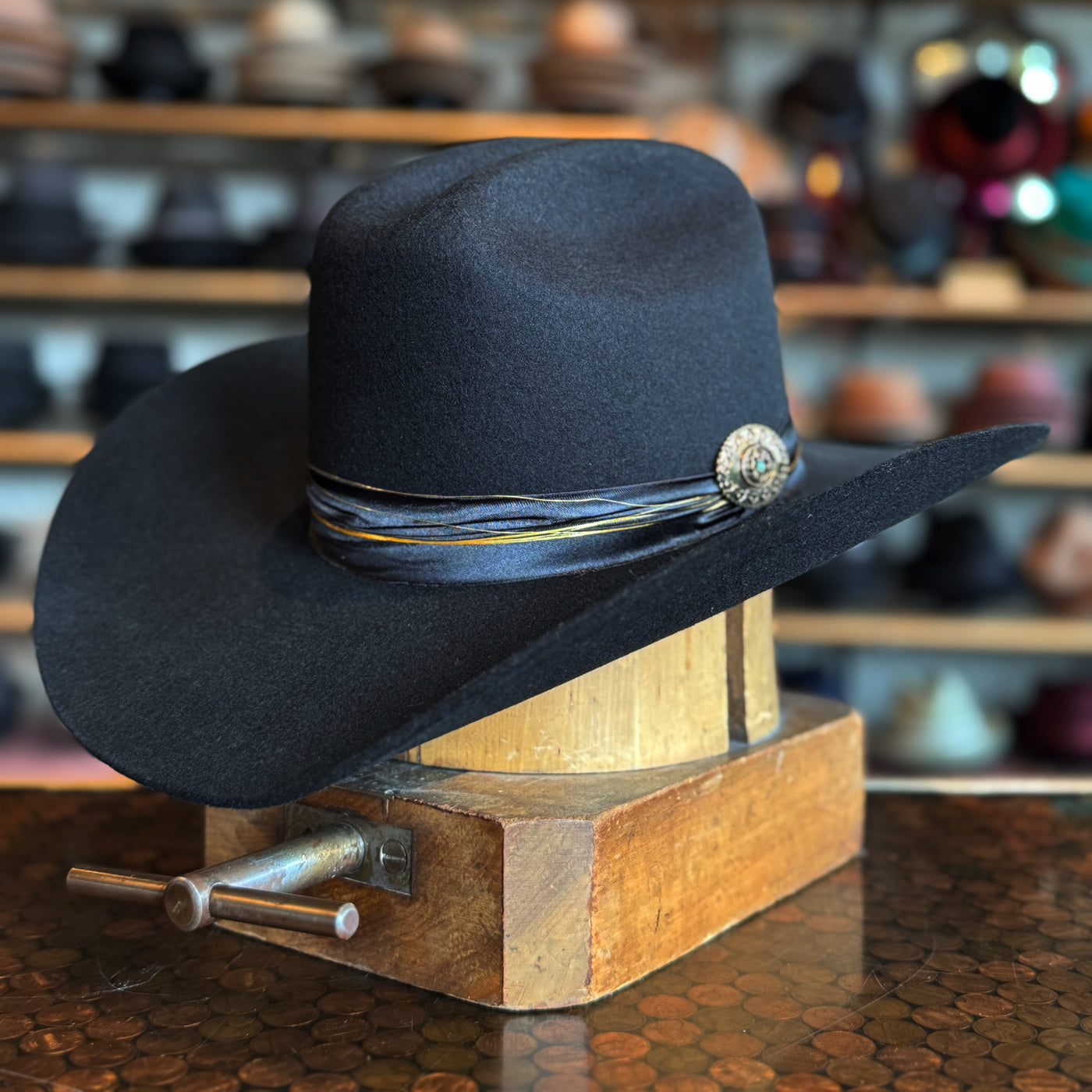 Daredevil black felt hat with wide brim, decorated with a thin striped hatband and metallic concho, displayed on a wooden hat block in a hat shop.