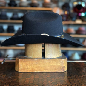 Daredevil black wide-brim hat resting on a wooden hat block with a leather band detail, displayed in a shop with various other hats blurred in the background.