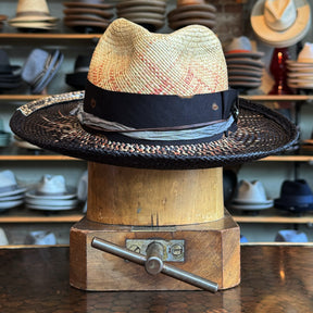 Dark Star Mel Fedora featuring a woven natural straw crown with subtle red accents, a wide black braided brim, and a layered ribbon band, displayed on a wooden hat block.