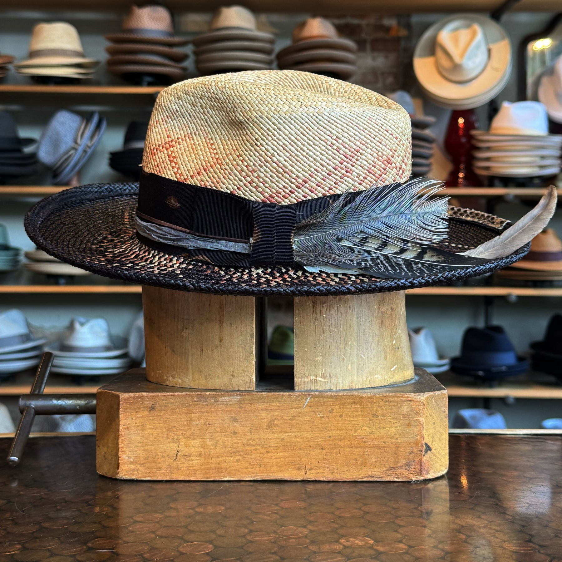 Dark Star Mel Fedora featuring a two-tone woven design with a natural crown and black brim, adorned with a black band and decorative feather, displayed on a wooden hat block.