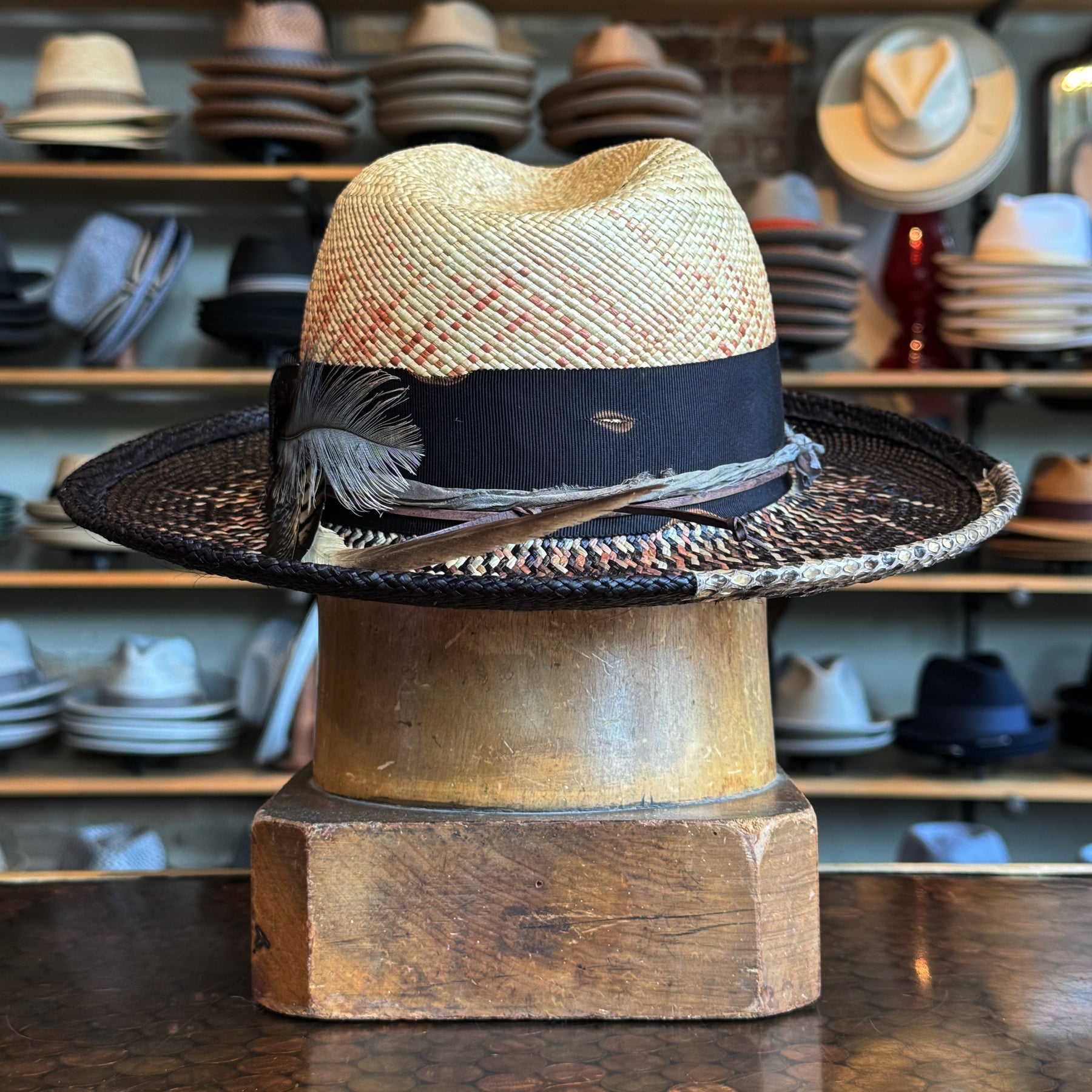 Dark Star Mel Fedora featuring a woven straw crown with a black wide brim, decorated with feathers and a black band, displayed on a wooden hat block in a hat shop.