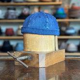 Denim Docker flat cap made from dark wash denim fabric displayed on a wooden hat block with a blurred background of various hats on shelves.