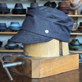 Denim Military Cap displayed on a wooden hat block, featuring a structured brim and classic military style in dark denim fabric, with various hats blurred in the background.