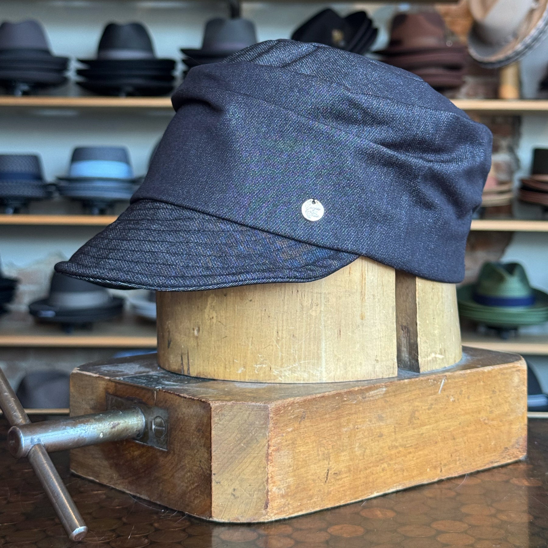 Denim Military Cap displayed on a wooden hat block, featuring a structured brim and classic military style in dark denim fabric, with various hats blurred in the background.