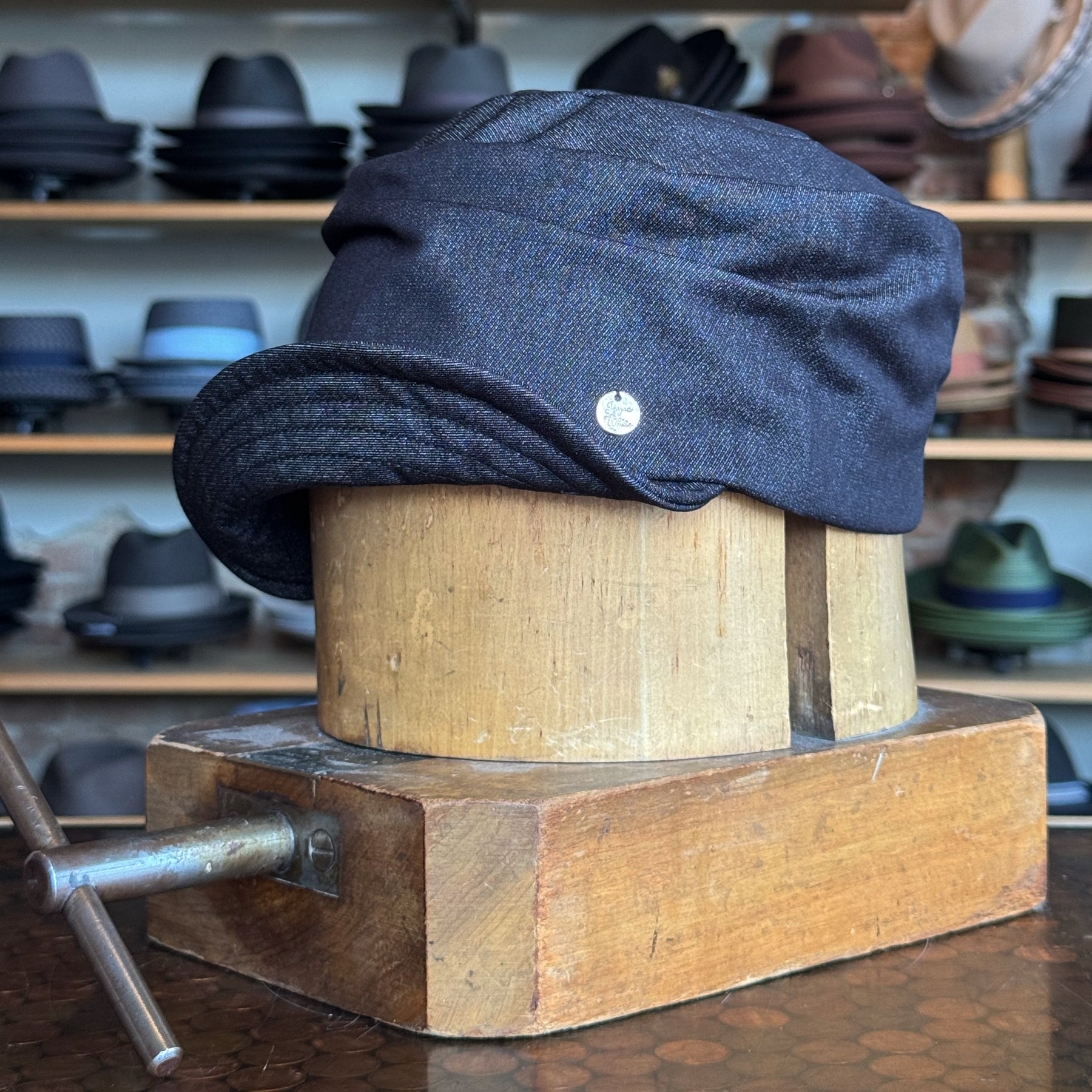 Denim Military Cap displayed on a wooden hat block, featuring a dark denim fabric with a rounded crown and a short curved brim, surrounded by various hats on shelves.