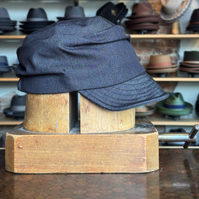 Denim Military Cap in dark blue fabric with a structured brim, displayed on a wooden hat block against a backdrop of assorted hats on shelves.