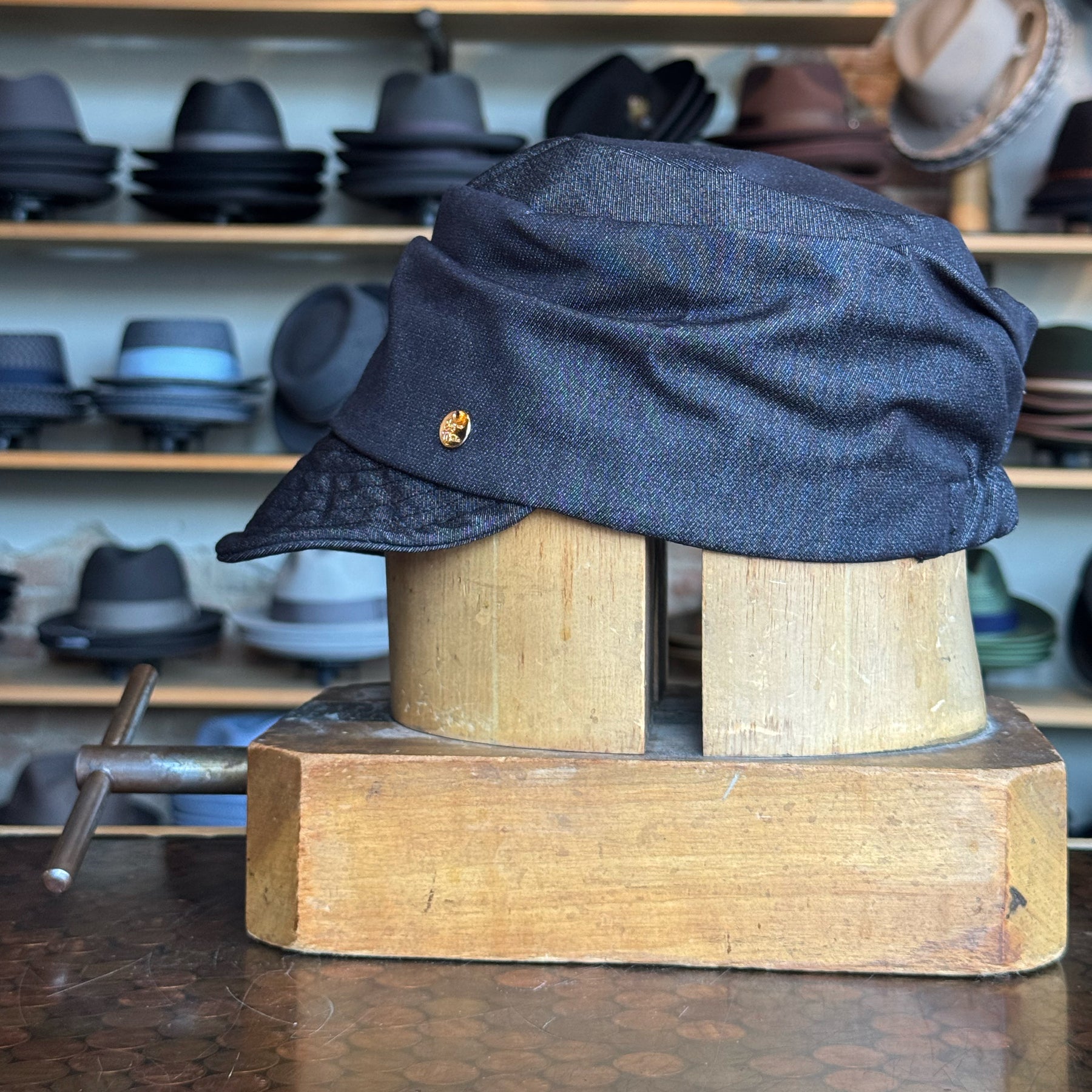Denim Military Cap in dark blue denim fabric displayed on a wooden hat block with multiple stylish hats in the background, showcasing a vintage-inspired accessory.